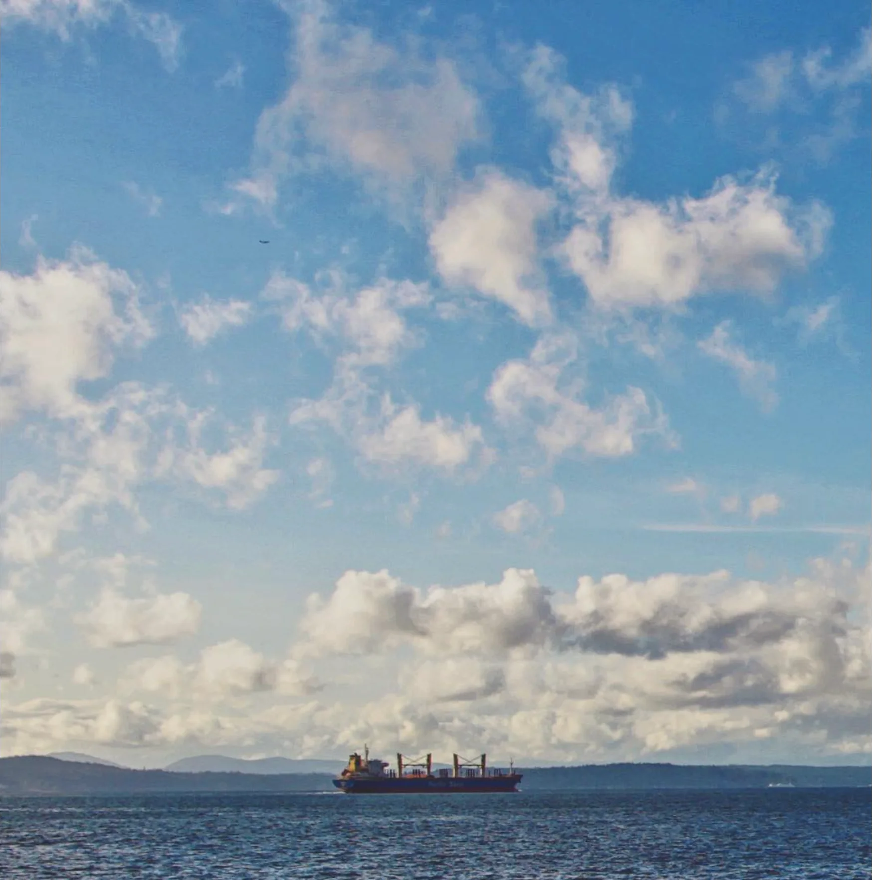 Cargo ship crossing open water beneath a broad blue sky filled with layered clouds.